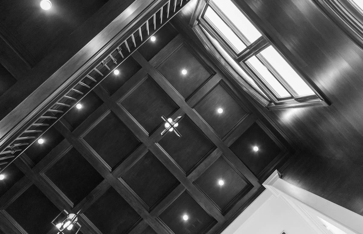 Black and white view looking up at a wooden coffered ceiling with recessed lights and a skylight.