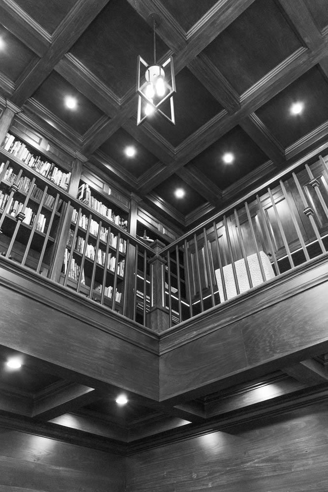 Two-story library with wood paneling, bookshelves, and a balcony. A geometric chandelier hangs from the coffered ceiling.