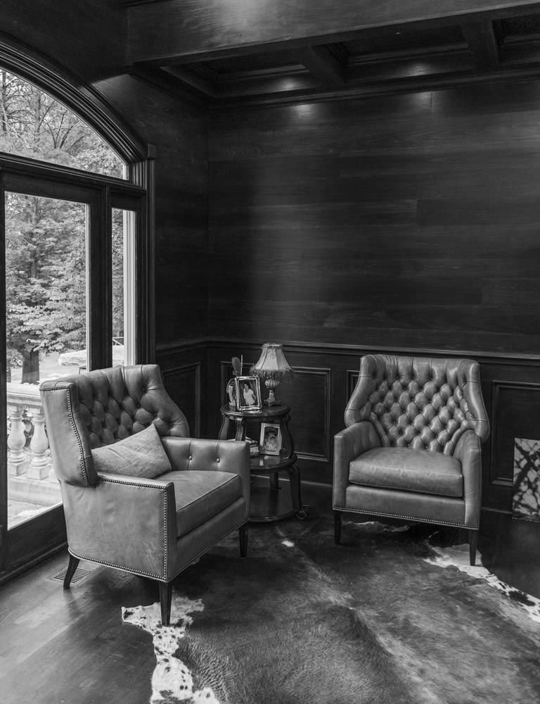 Two tufted armchairs in a dark-paneled room with a cowhide rug and a view of trees through a large window.
