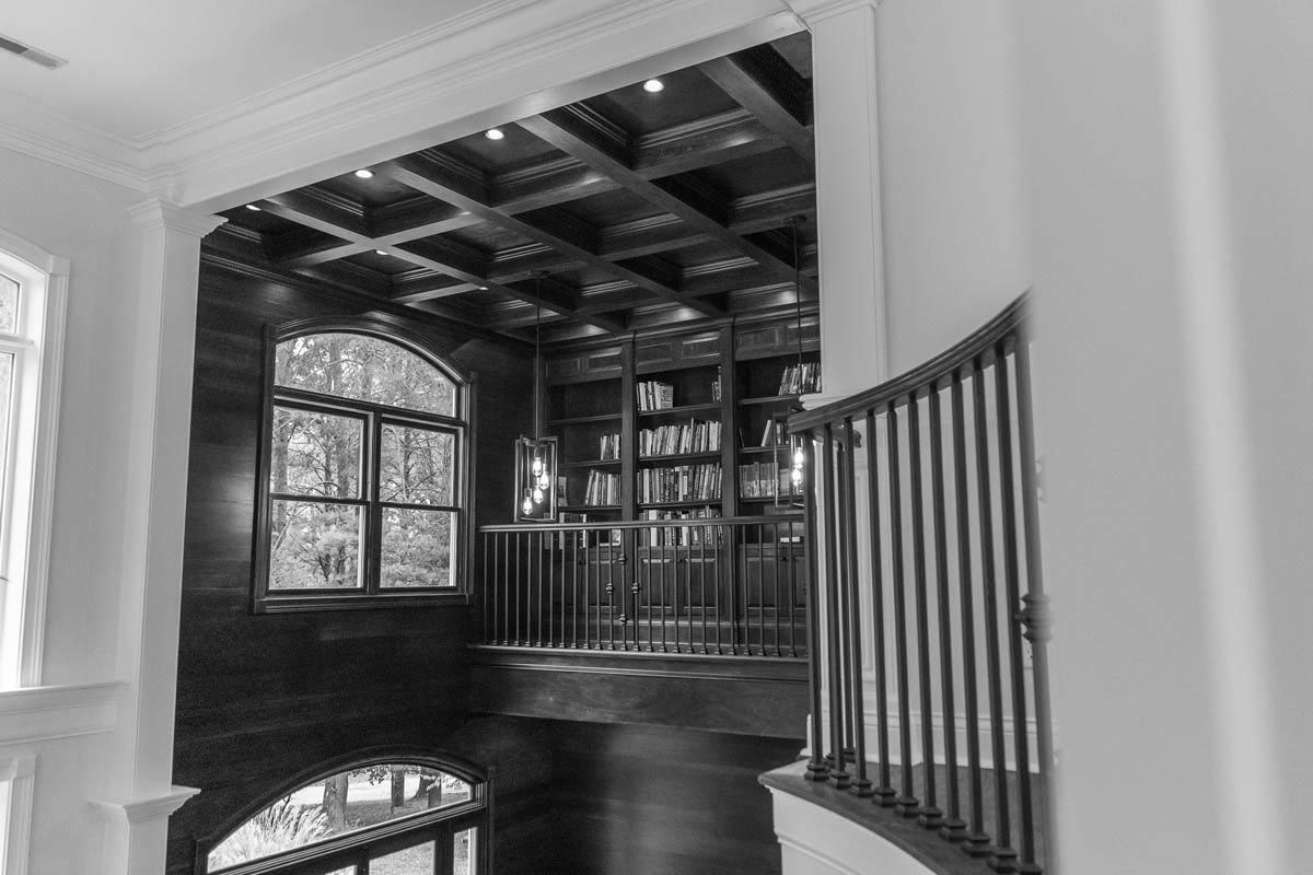 Library with wood-paneled walls, arched window, and coffered ceiling. Bookshelves, a railing, and dark flooring.