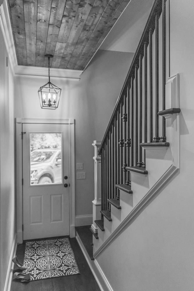 Black and white image of a hallway with a wood ceiling, black rod chandelier and staircase.