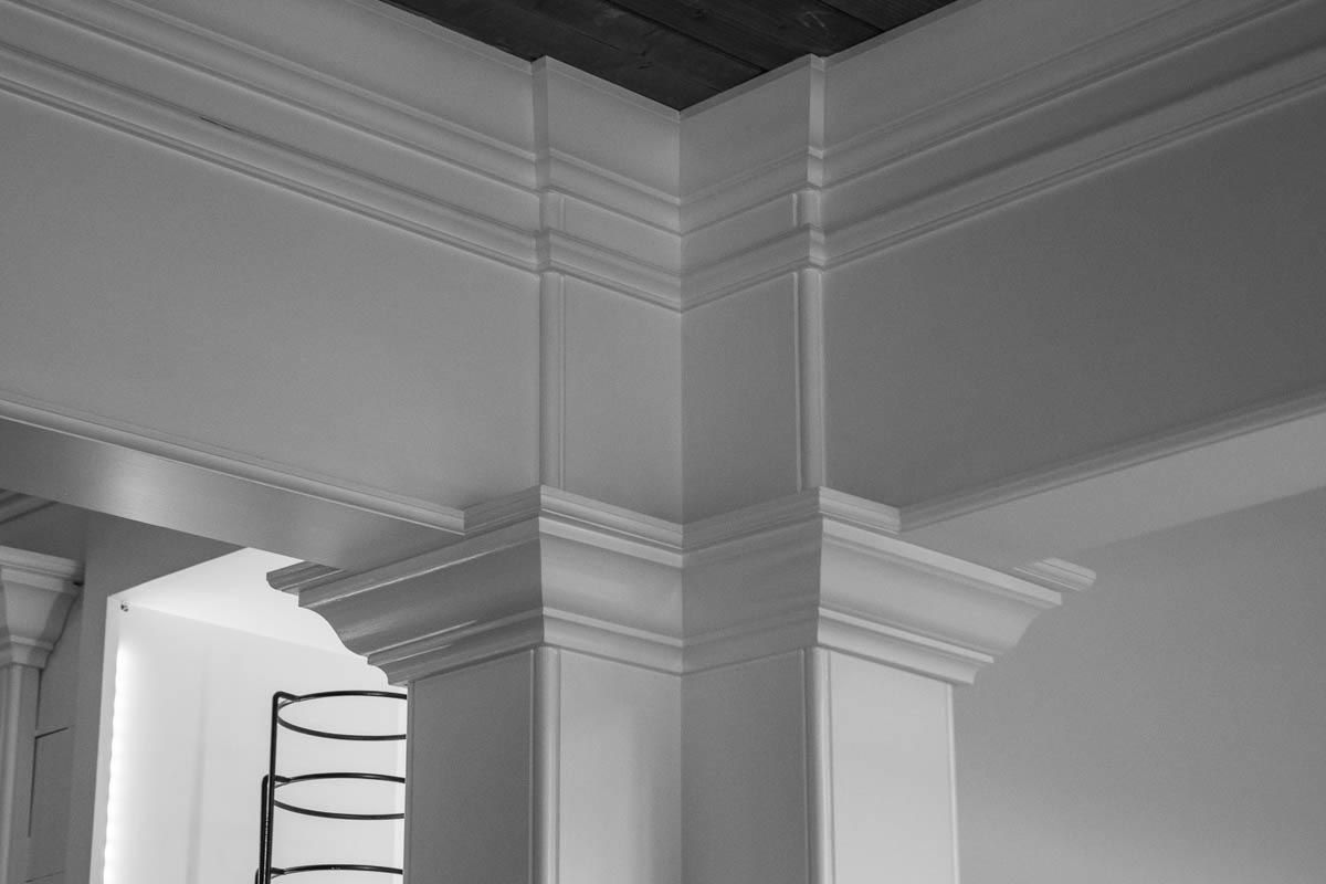 Corner of a white, decorative interior with detailed crown molding around a column and ceiling.