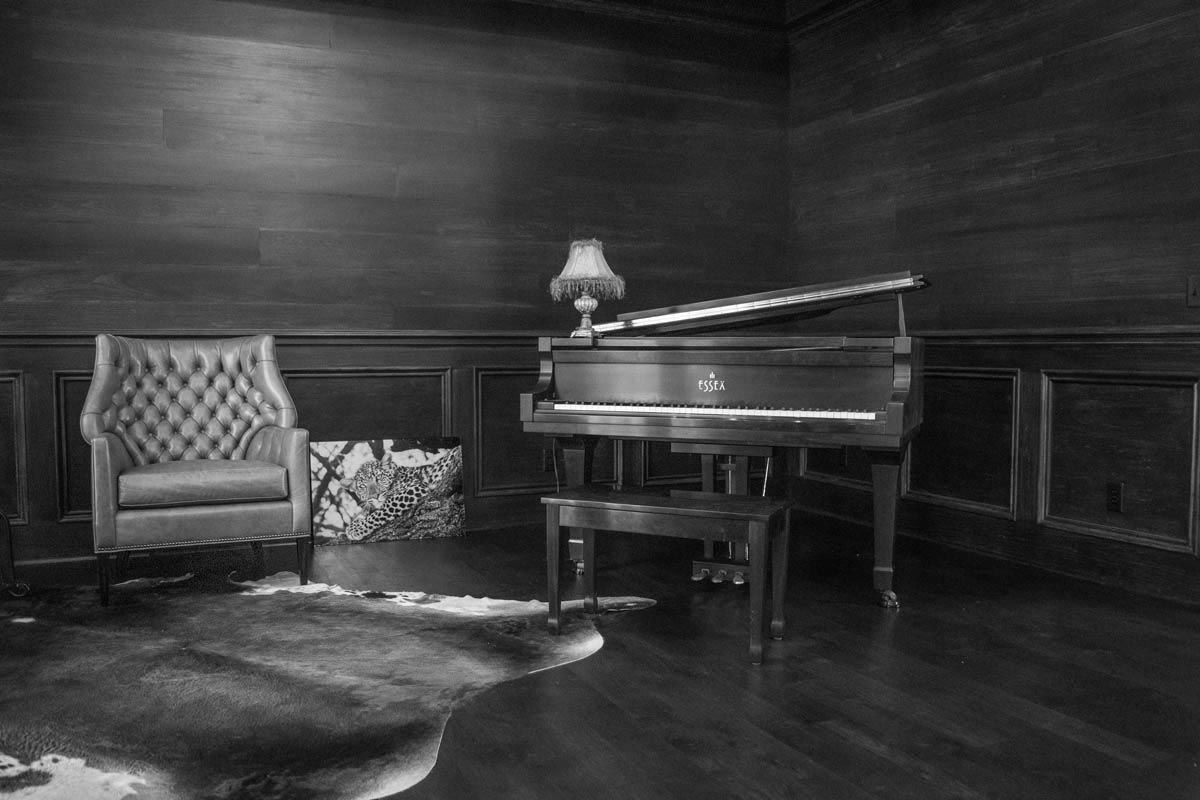Black and white image: A grand piano, chair, and lamp in a wood-paneled room on a patterned rug.