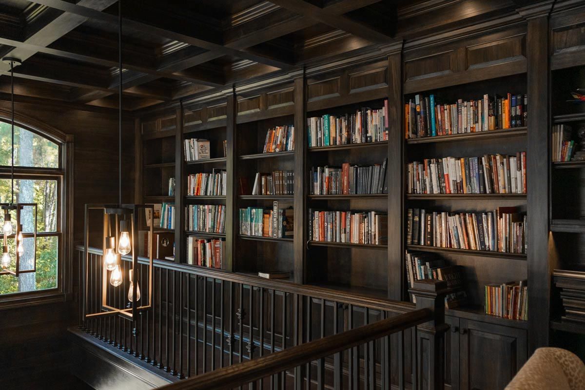 Dark wood library with built-in bookshelves, railing, and a window overlooking greenery.