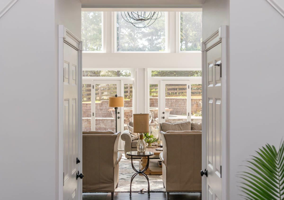 View through open white doors into a living room with tan furniture and large windows.