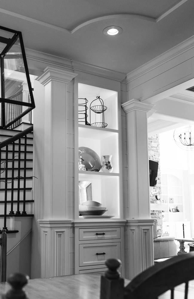 Built-in white cabinet with open shelving displaying decorative items, next to a staircase and column in a home.