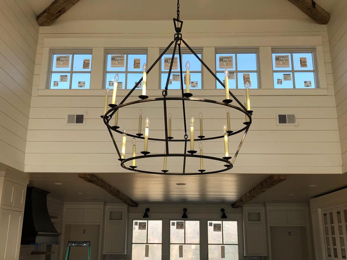 Large chandelier hangs in a high-ceilinged room with windows. White walls, wood beams.