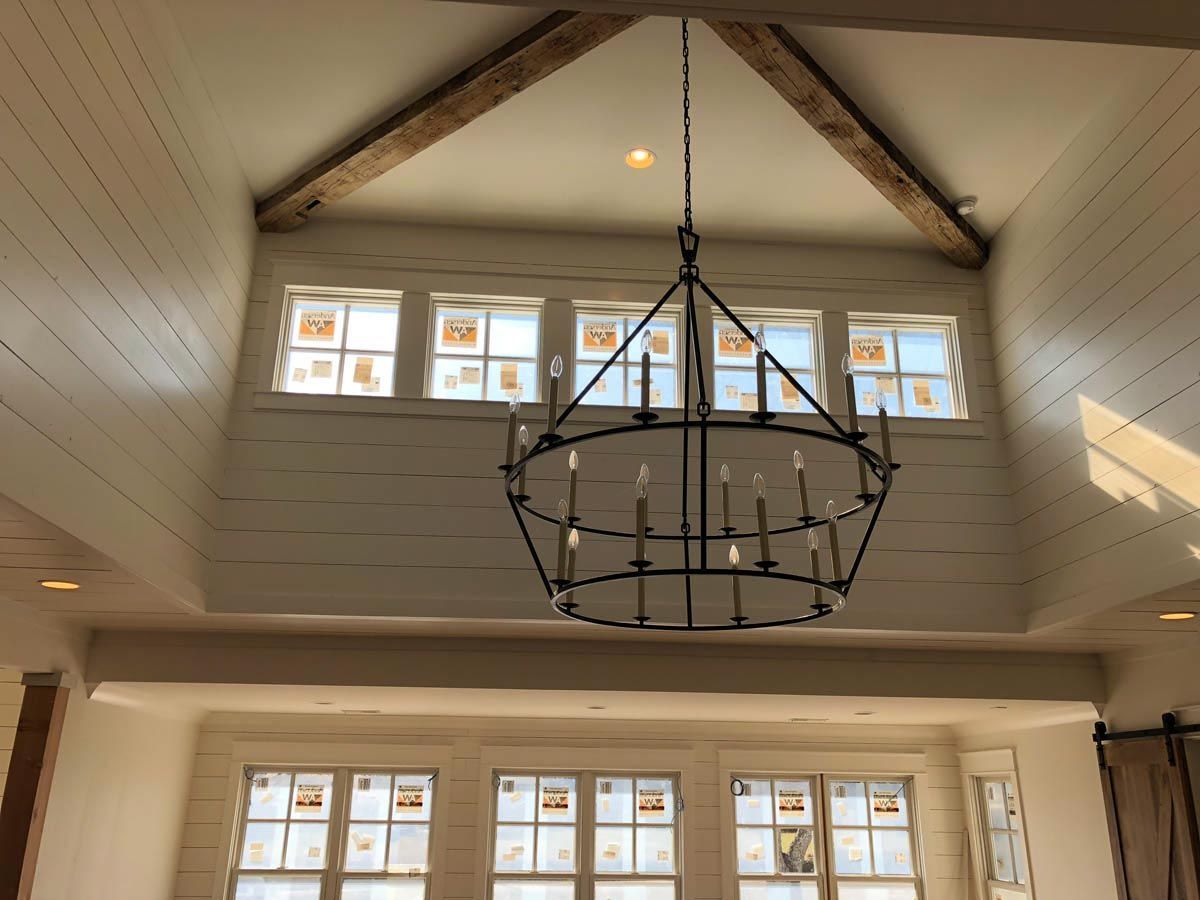 An ornate chandelier hangs in a room with white paneled walls and windows, illuminated by sunlight.