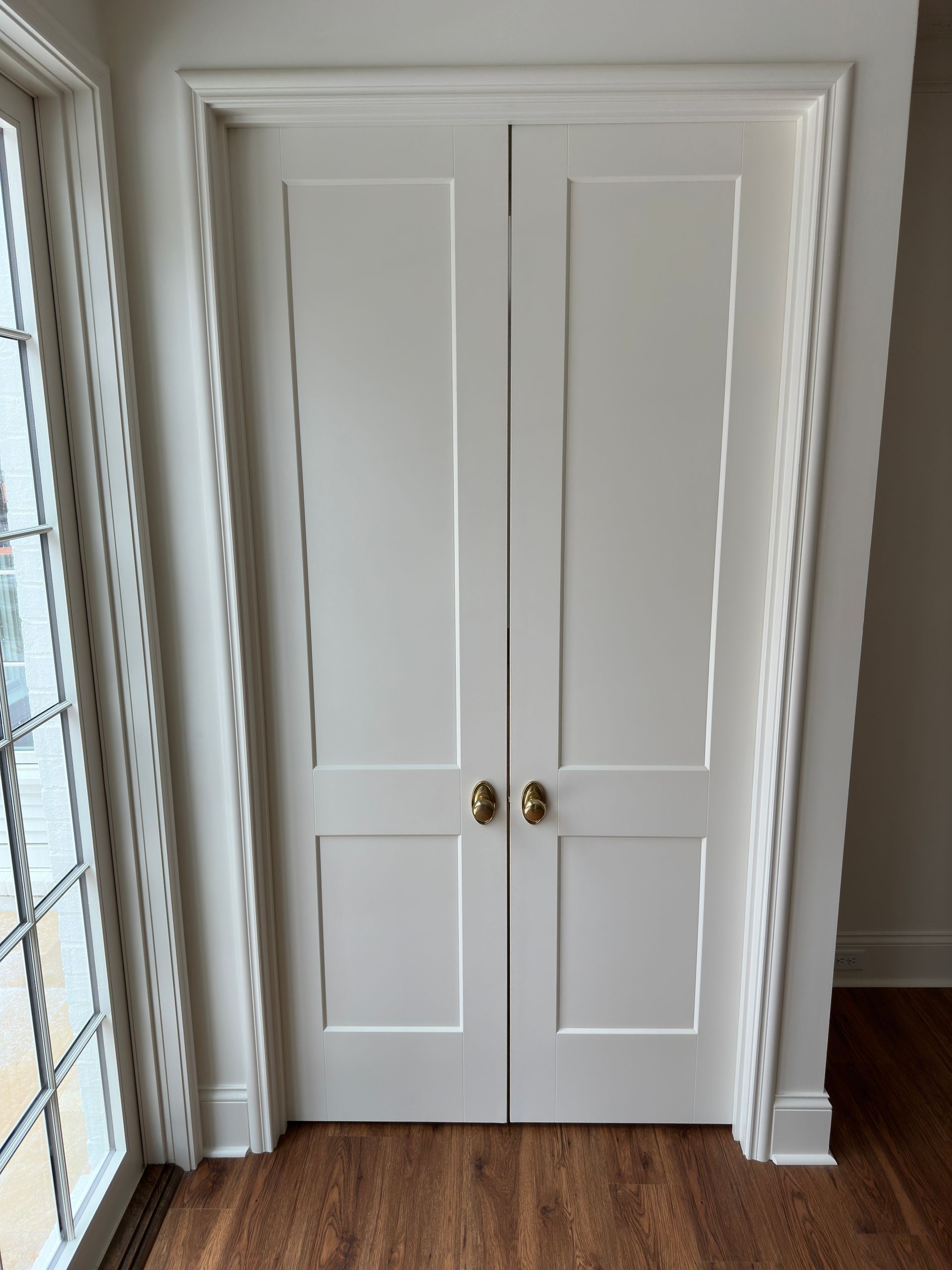 White, two-panel, double doors with gold hardware; located between a window and a wall.