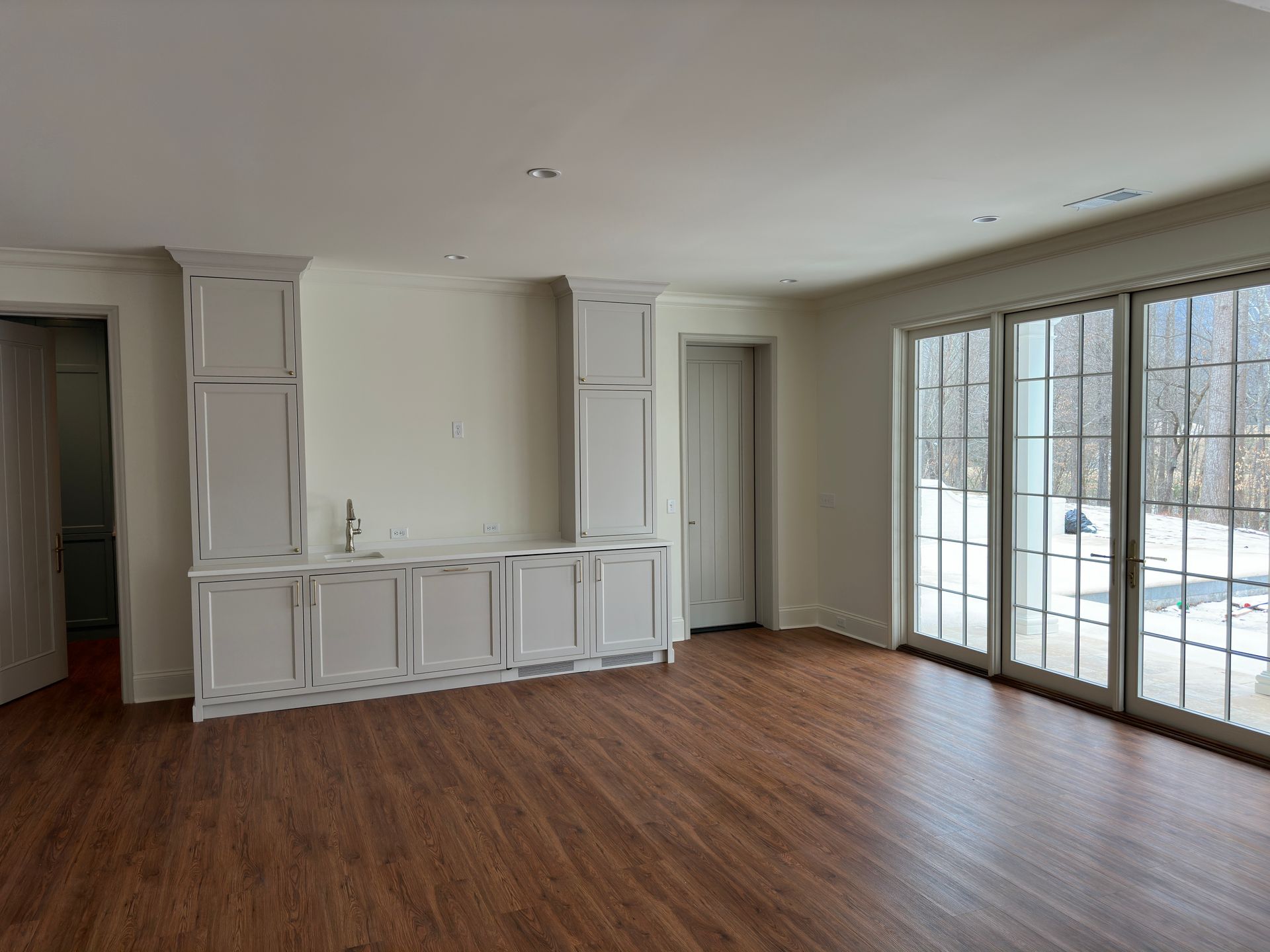Empty room with built-in white cabinets and dark wood-look flooring. Large glass doors and a door are visible.