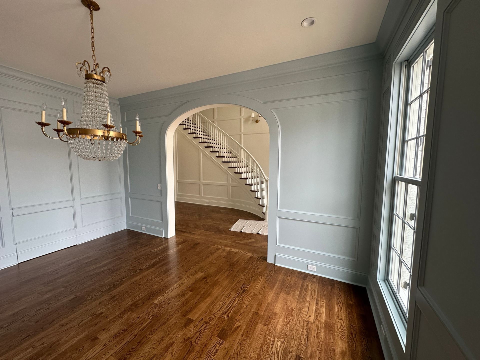 Formal dining room with wood floors, blue-gray walls, chandelier, arched doorway leading to a staircase, and a window.