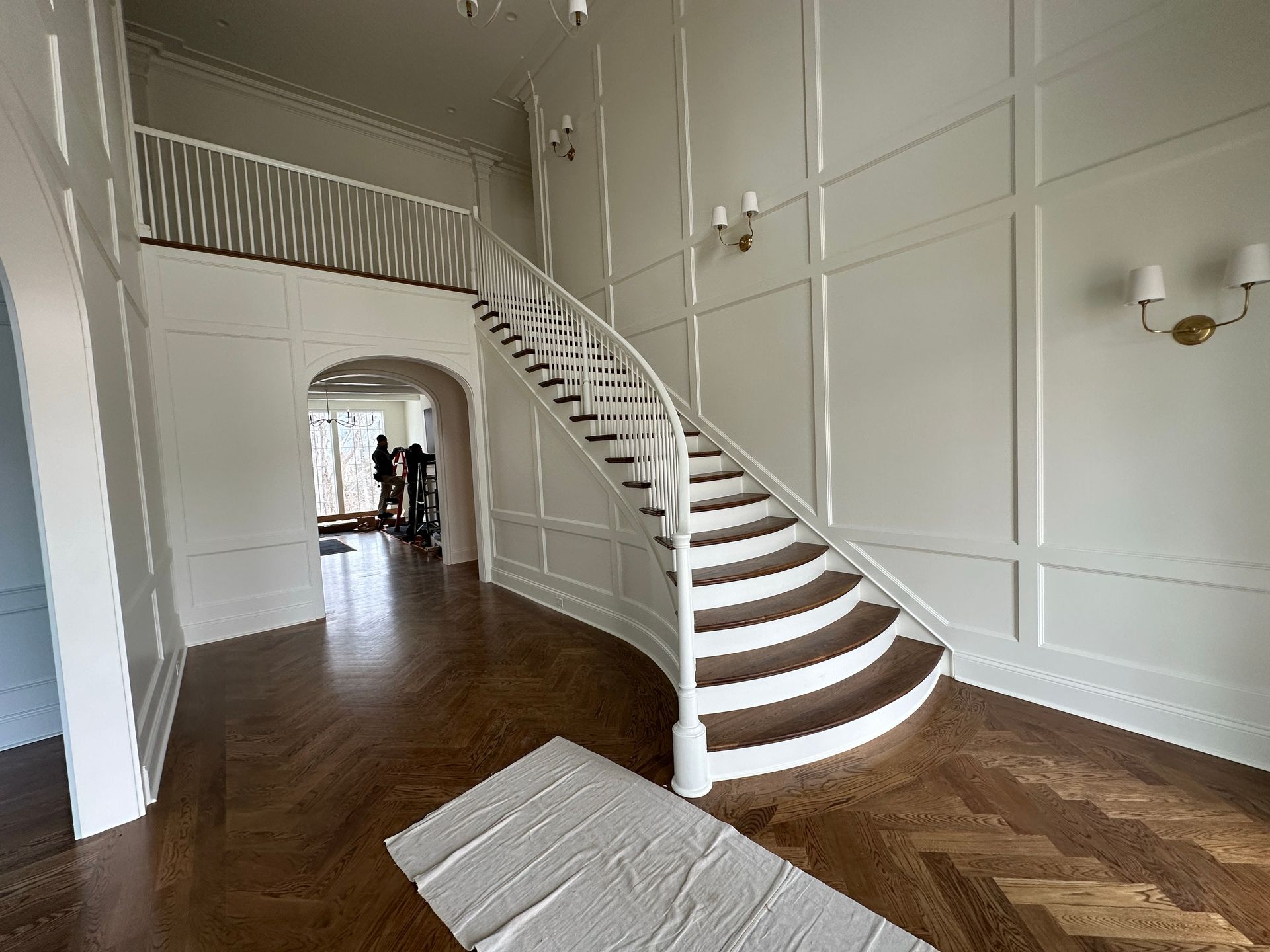 White staircase in a large, bright room with hardwood floors. Paneling on the walls, and a protective cloth on the floor.