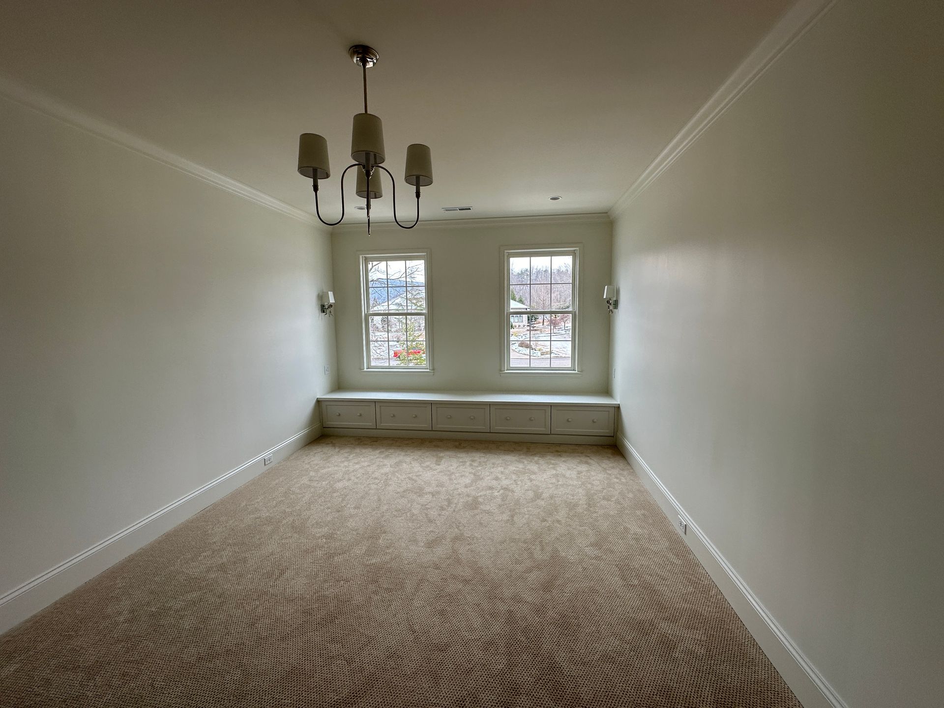 Empty room with patterned carpet, two windows with built-in bench, chandelier. White walls.