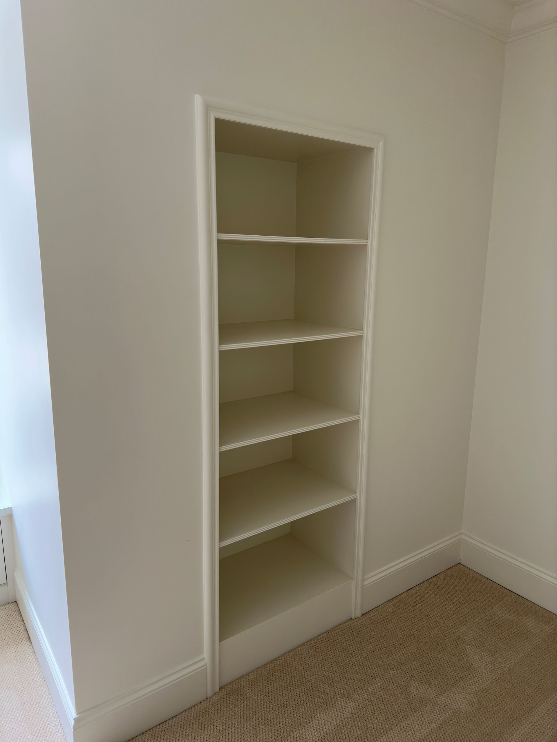 Built-in white bookshelf in a wall niche; beige carpet and white walls.