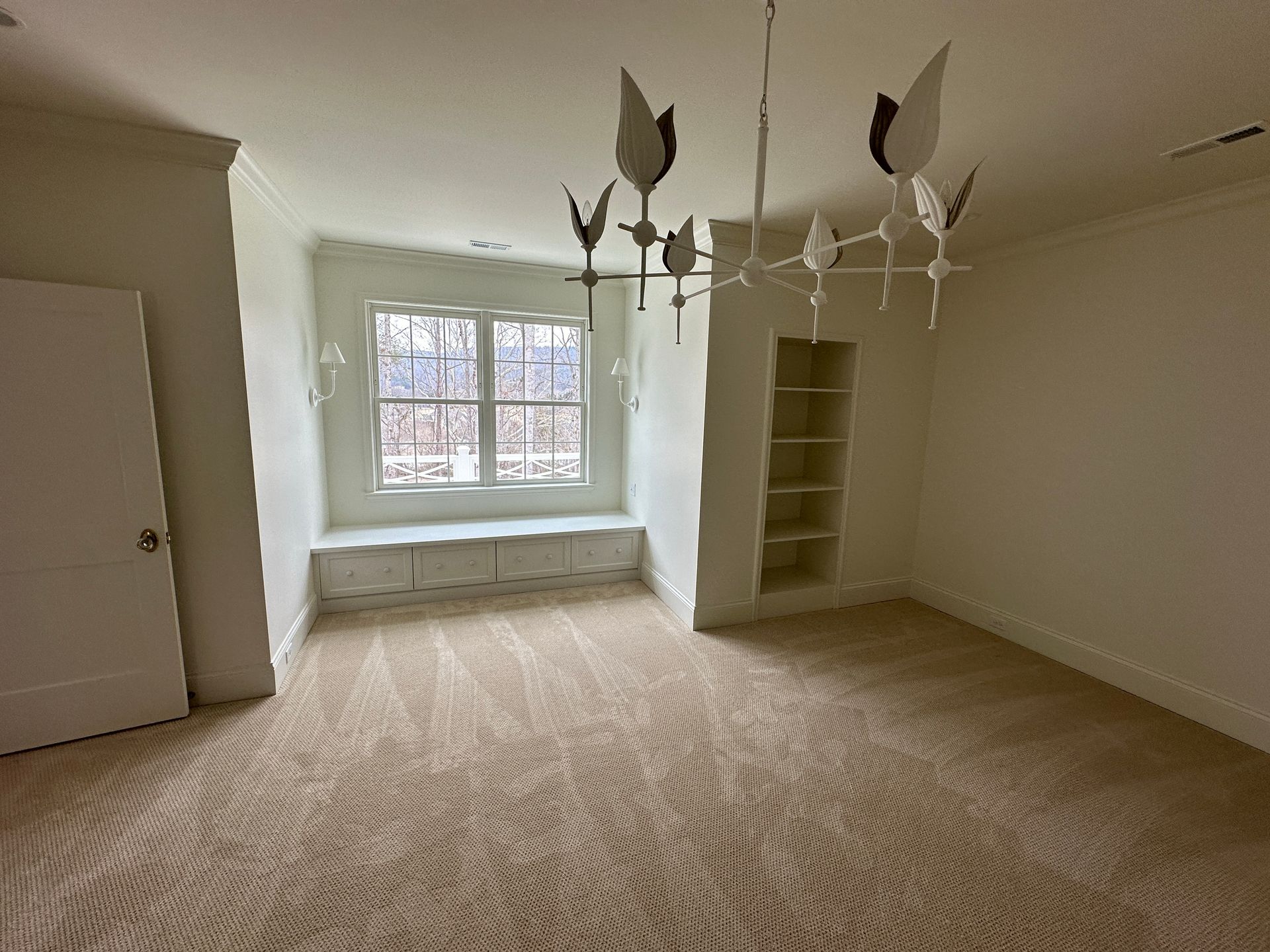 Empty room with beige patterned carpet, white walls, built-in window seat, and unique chandelier.