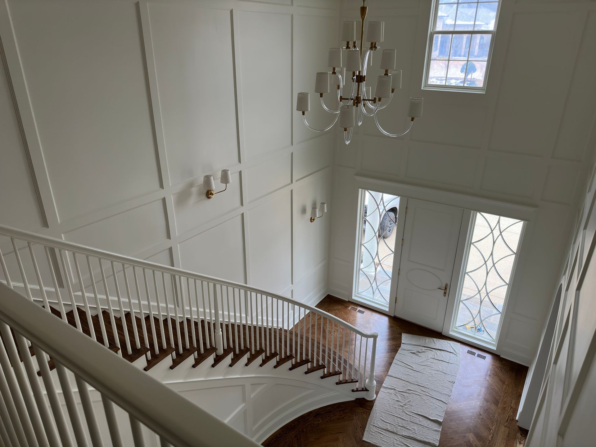 Grand white entryway with staircase and chandelier. White paneled walls and dark wood floor.