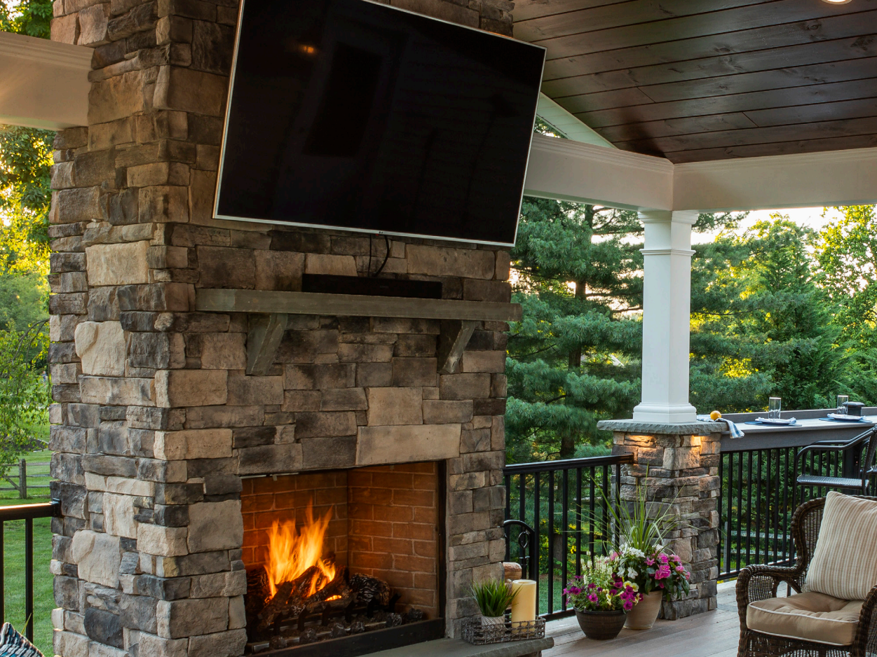 Stone fireplace with burning fire; mounted TV above. Outdoor patio setting with greenery.
