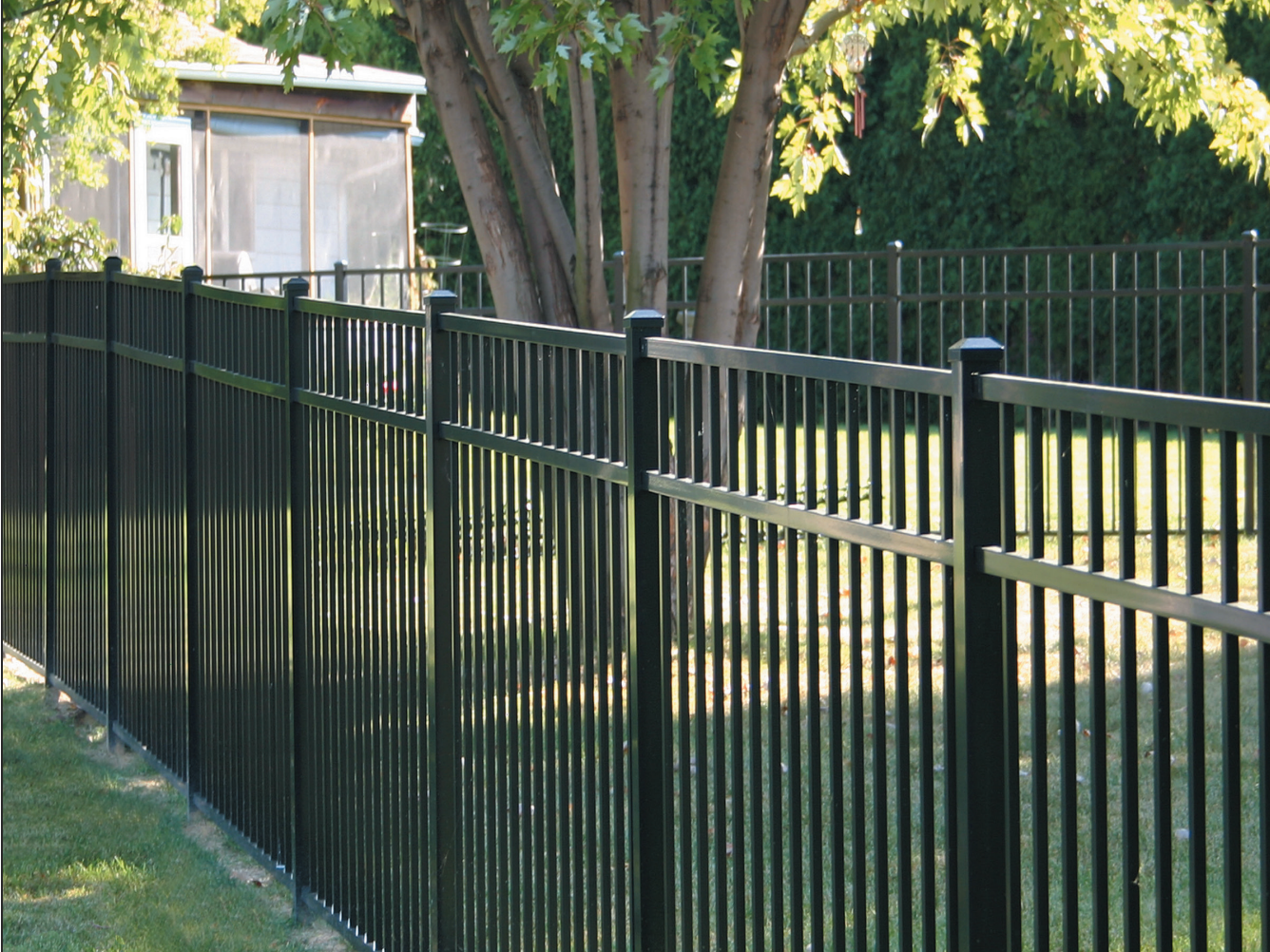 Black metal fence encloses a green lawn, with a tree and small building visible in the background.