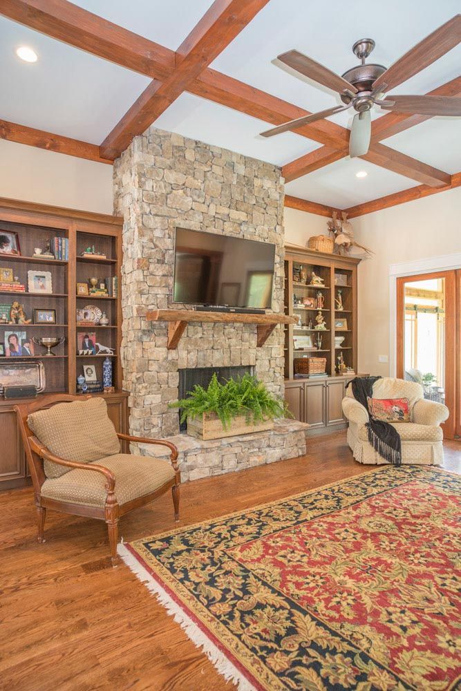 Living room with a large stone fireplace with a mounted tv, wood beams and a ceiling fan.