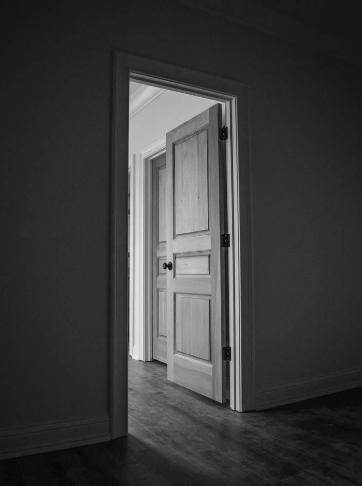 Black and white photo of a partially open door, revealing another open door in a room with hardwood floors.