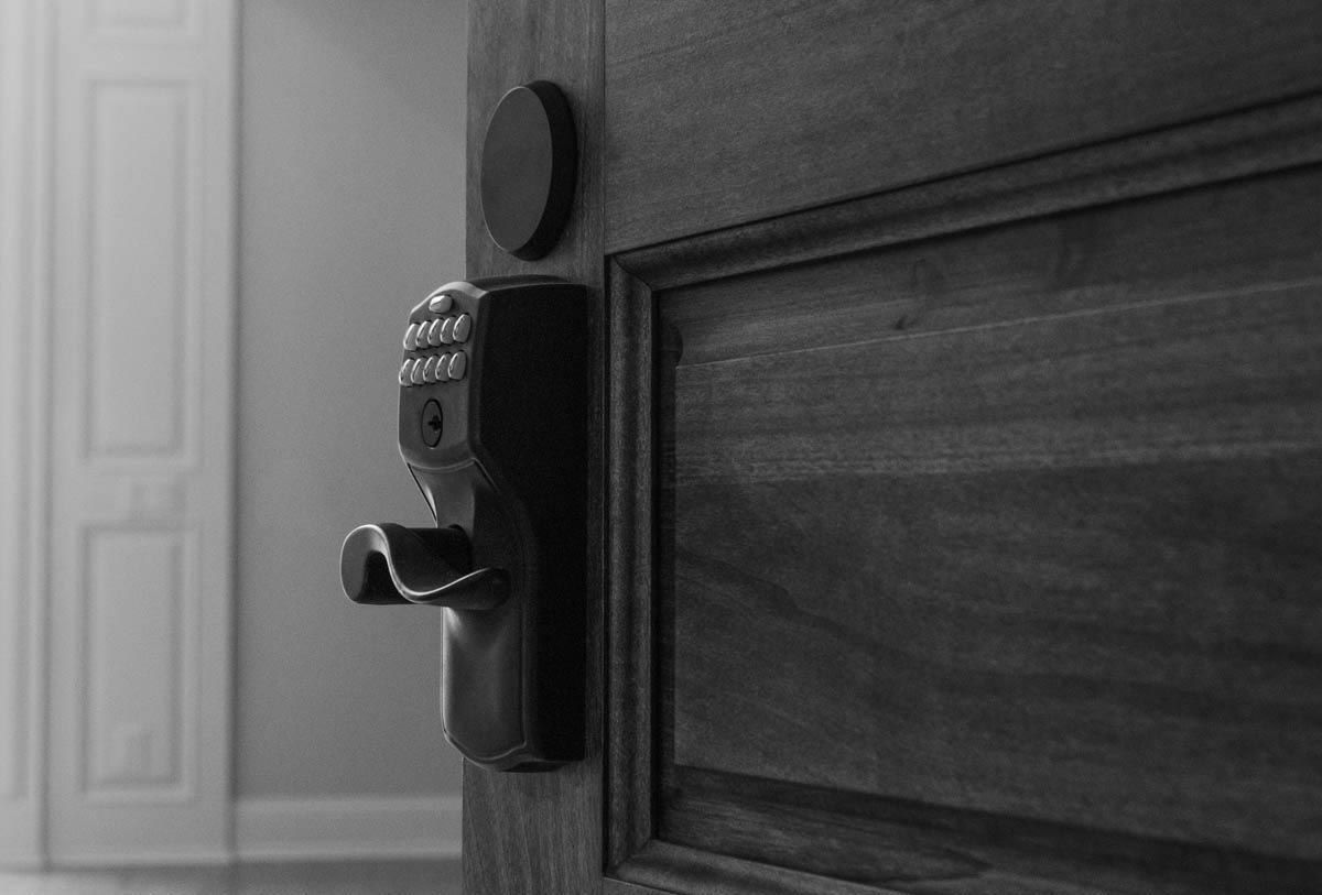 Black and white close-up of a dark wooden door with a smart lock, slightly ajar, revealing a hallway.