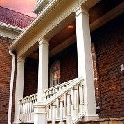 White-columned porch with stairs, railing, and a window; red brick wall.