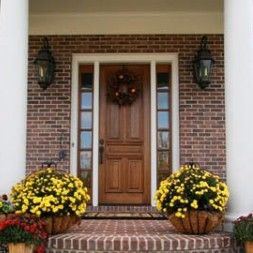 A brick house entrance with a wooden door, two potted yellow flowers, and wall lanterns.