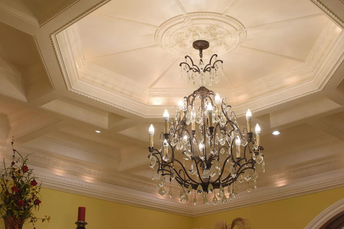Ornate chandelier hangs from an octagon-shaped ceiling with detailed molding. Warm lighting in a luxurious interior.