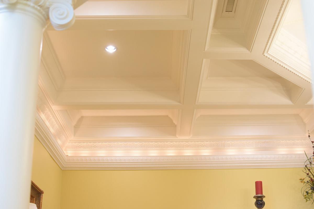 Cream-colored coffered ceiling with ornate trim, a light fixture, and a partial view of a white column.