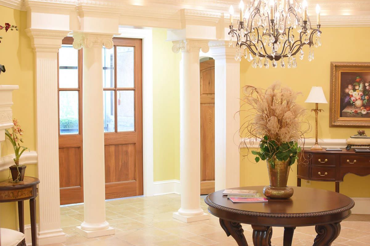 Elegant foyer with columns, wood door, chandelier, and round table. Yellow walls, floral arrangement.