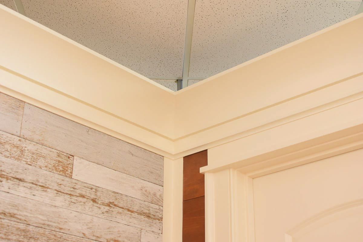 Cream-colored crown molding at a corner, with textured wood-look wall and white door frame. Beige ceiling.