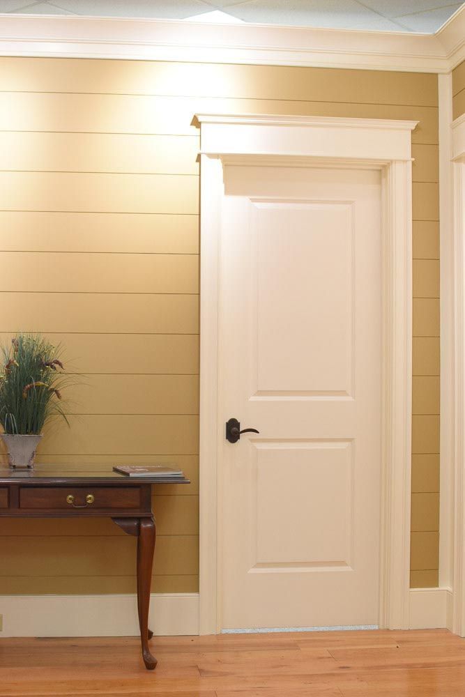 White door with black handle, in off-white trim against a yellow horizontal plank wall, side table with plant.