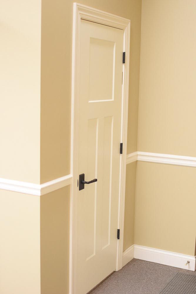 Narrow, white door with black hardware in a corner of tan walls with white trim.