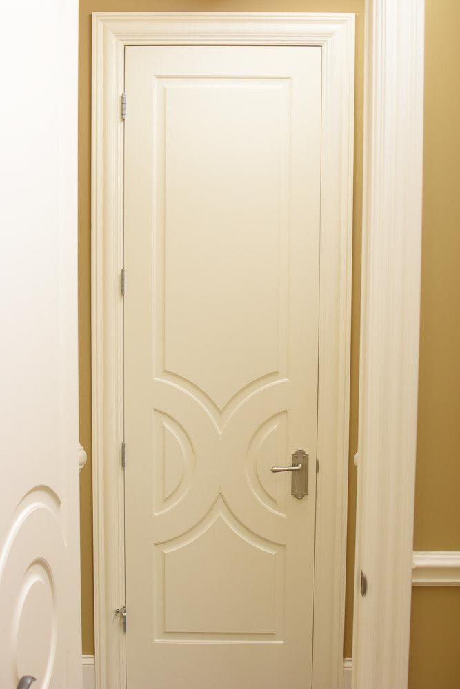 White paneled door with decorative design, silver handle, set in a cream-colored doorway and against a tan wall.
