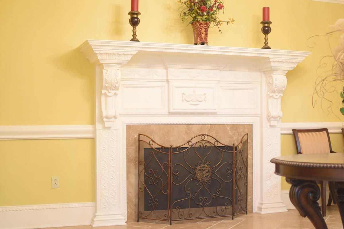 White ornate fireplace with a black screen on a yellow wall, with candles and a floral arrangement on the mantel.