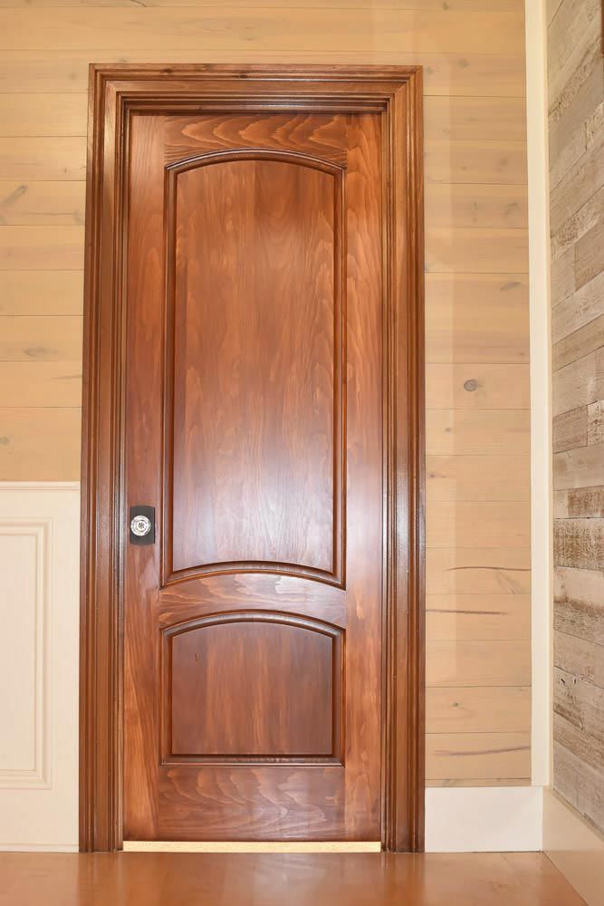 Wooden door with arched panels and dark hardware in a light wood-paneled wall.