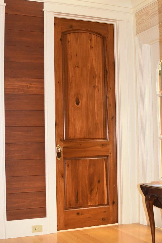 Wooden door with arched top panel and rectangular bottom panel, framed by white molding, near a wood-paneled wall.