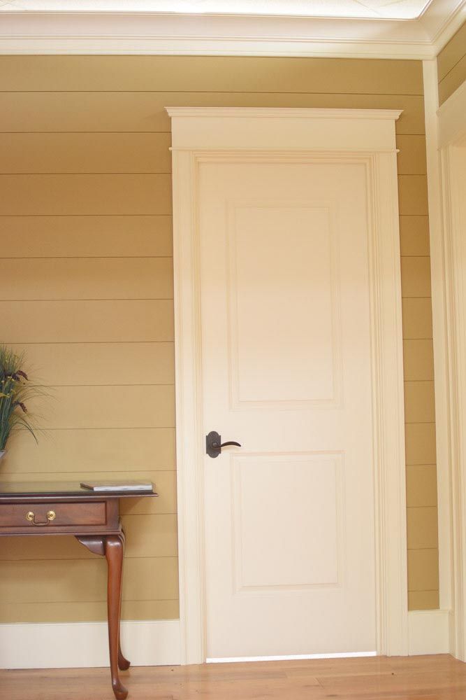 Cream-colored door with black handle, framed by cream trim on a tan shiplap wall.