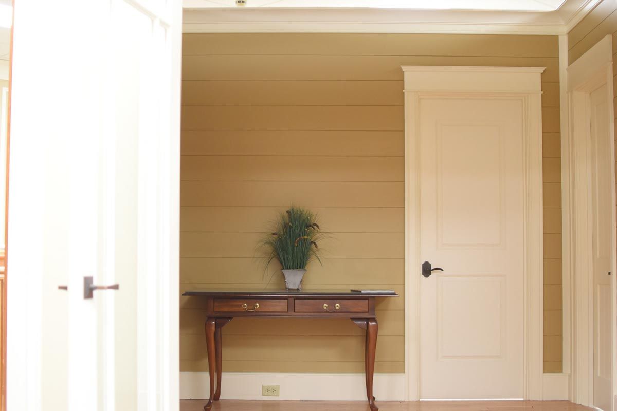 Interior hallway with a table, plant, and a closed door. Light wood walls and trim.