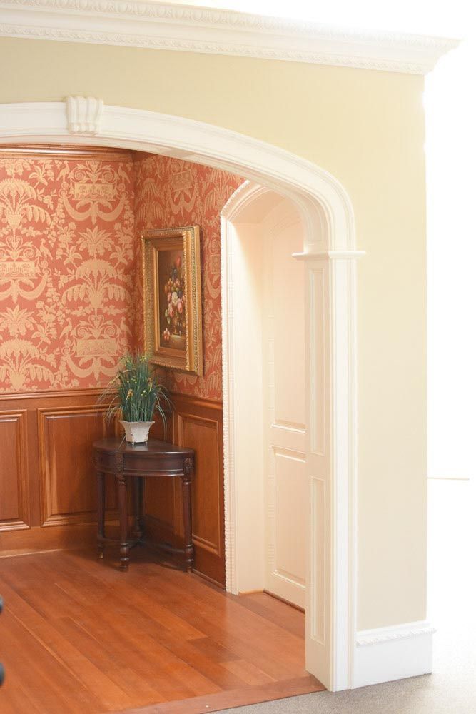 Archway leading to a room with red patterned wallpaper, wooden table, painting, and potted plant.