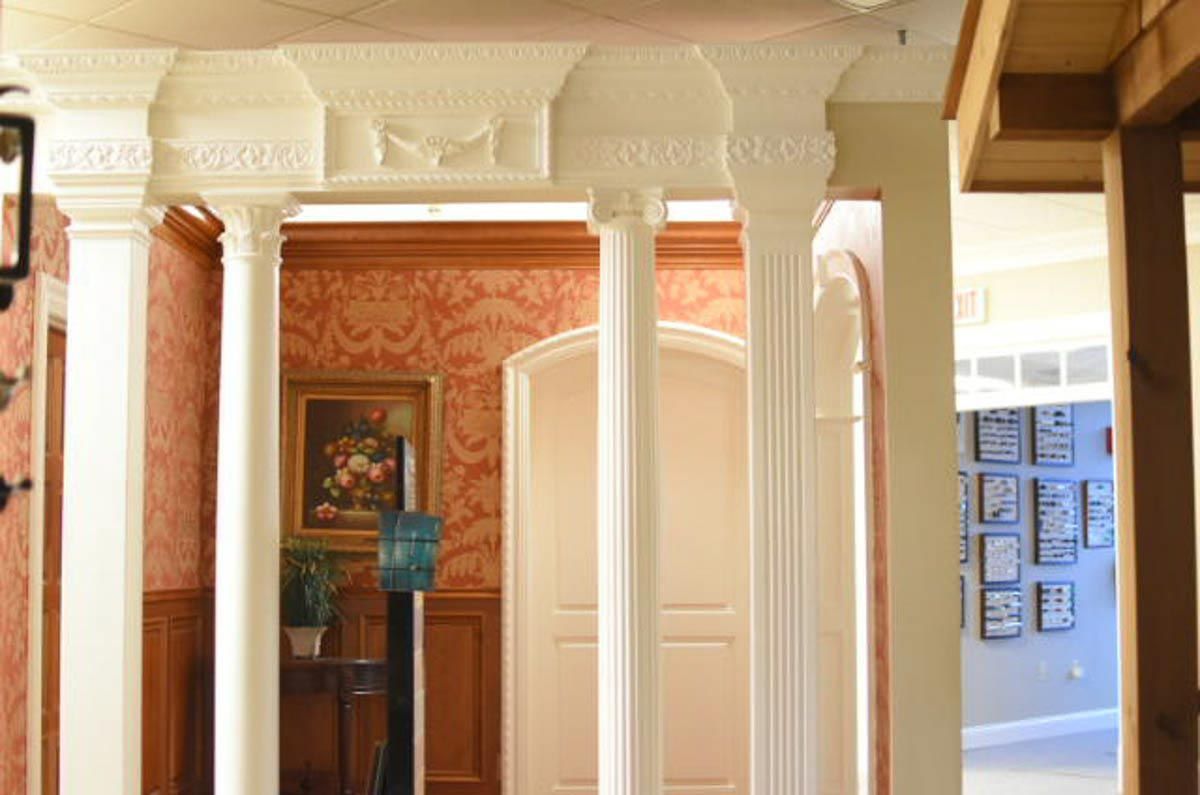 White columns with ornate capitals frame a doorway; red patterned wallpaper and a painting are visible.