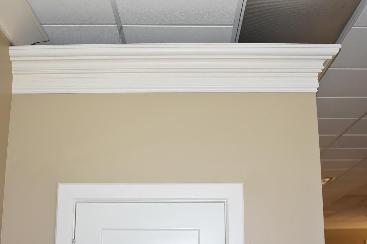 White crown molding above a white doorframe on a beige wall, a suspended ceiling is in the background.