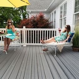 Two women on a deck with drinks, one under a yellow umbrella. White railing, gray deck, lush green background.
