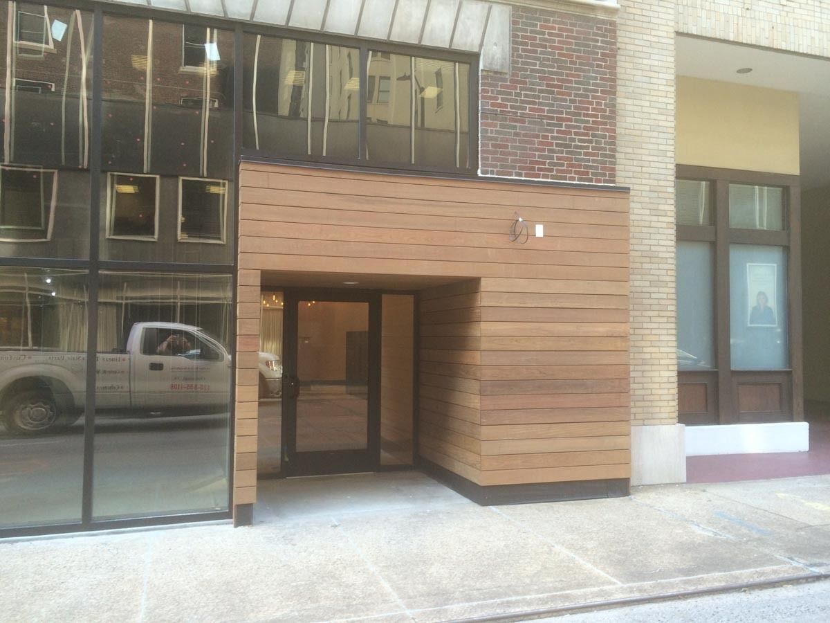 Entrance to a building with glass windows, wood paneling, and a brick facade. A white truck is visible in the reflection.