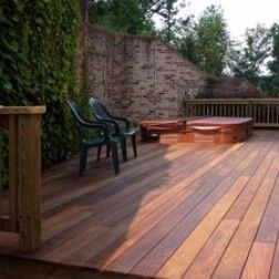Wooden deck with a hot tub and two chairs, against a brick wall, shaded by trees.