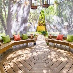 Wooden gazebo with seating, cushions, and sheer curtains, in a sunlit garden.