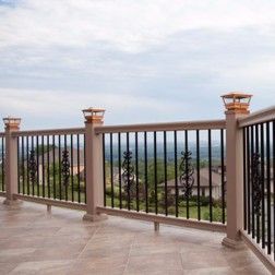 Tan deck railing with black vertical bars and decorative accents, overlooking a distant landscape under a cloudy sky.