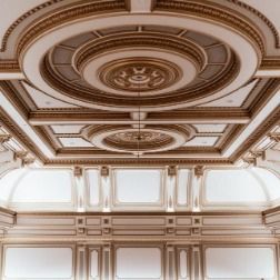 Ornate, gold-trimmed ceiling with circular and rectangular patterns. White walls with framed panels.