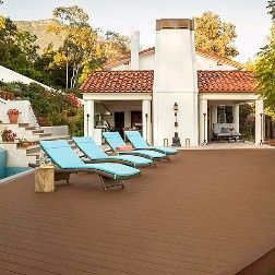 Lounge chairs on a brown deck in front of a white house with a terracotta roof, near a pool.