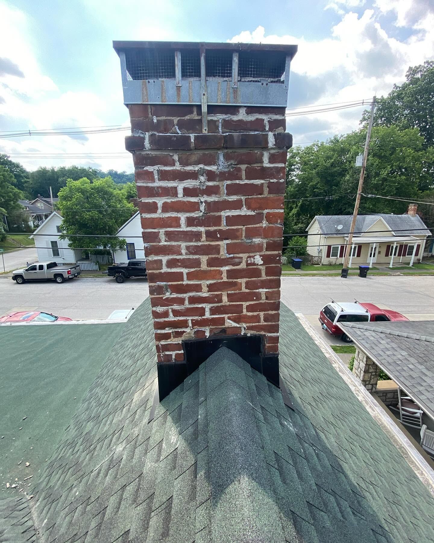 Brick chimney on a green shingled roof, with neighboring houses and a street in the background.